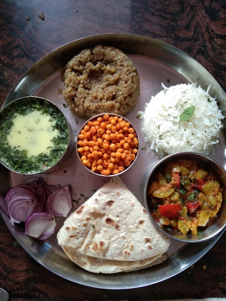 A traditional Indian meal served on a metal thali, featuring a variety of dishes. There is a portion of plain white rice, a serving of thick wheat-based porridge, and a flatbread. Accompaniments include a bowl of green curry, small crispy orange snacks, and a dish of colorful mixed vegetables. The meal is garnished with sliced red onions and fresh herbs.