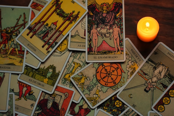 A variety of tarot cards are spread out on a wooden surface next to a lit candle. The cards show intricate illustrations and symbols, including figures and abstract designs, suggesting themes of divination or fortune-telling.