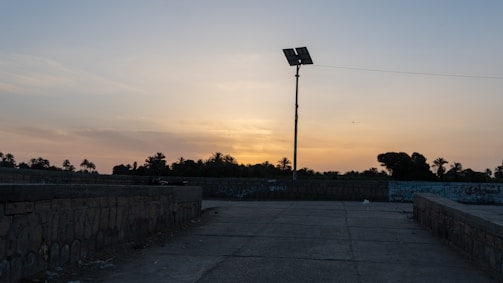 The scene features a sunset or early evening sky with a mix of light orange and blue colors. In the foreground, there is a paved area lined with stone walls covered in graffiti. A tall street lamp with solar panels is prominently visible, while palm trees and shrubbery are seen in the background, silhouetted against the horizon.