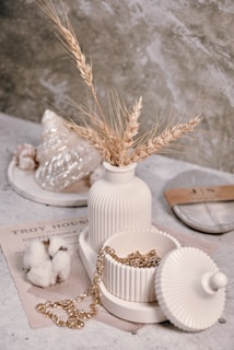 A ribbed ceramic vase holds dried wheat stalks, placed beside a matching lidded container with a gold chain spilling out. In the background, a translucent decorative shell and cotton branches rest on top of a vintage newspaper. The scene exudes a rustic and elegant aesthetic against a textured stone backdrop.