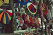 Colorful handcrafted textiles, including knitted hats and patterned bags, hang from a wooden stand. The variety of colors and patterns creates a vibrant display of traditional craft. In the blurred background, there appears to be a person wearing a yellow and blue scarf.