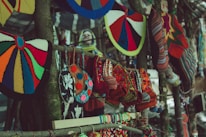 Colorful handcrafted textiles, including knitted hats and patterned bags, hang from a wooden stand. The variety of colors and patterns creates a vibrant display of traditional craft. In the blurred background, there appears to be a person wearing a yellow and blue scarf.