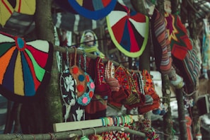 Colorful handcrafted textiles, including knitted hats and patterned bags, hang from a wooden stand. The variety of colors and patterns creates a vibrant display of traditional craft. In the blurred background, there appears to be a person wearing a yellow and blue scarf.