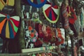 Colorful handcrafted textiles, including knitted hats and patterned bags, hang from a wooden stand. The variety of colors and patterns creates a vibrant display of traditional craft. In the blurred background, there appears to be a person wearing a yellow and blue scarf.