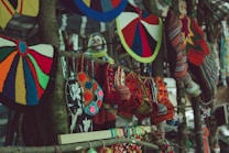 Colorful handcrafted textiles, including knitted hats and patterned bags, hang from a wooden stand. The variety of colors and patterns creates a vibrant display of traditional craft. In the blurred background, there appears to be a person wearing a yellow and blue scarf.