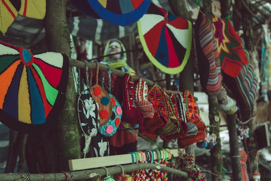 Colorful handcrafted textiles, including knitted hats and patterned bags, hang from a wooden stand. The variety of colors and patterns creates a vibrant display of traditional craft. In the blurred background, there appears to be a person wearing a yellow and blue scarf.