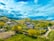 A small, rural village nestled in lush hills under a vibrant blue sky dotted with clouds. The landscape consists of scattered houses with reddish-brown rooftops surrounded by abundant greenery and winding dirt paths. Rolling hills and power lines are visible in the distance, creating a serene and picturesque setting.