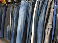 A row of jeans in various shades of blue and black is displayed on hangers in a clothing store. The jeans are arranged closely together, demonstrating a variety of washes and styles, from light blue to dark denim.