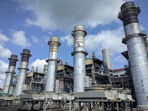 Several large industrial chimneys tower over a complex network of metal structures and machinery, set against a partly cloudy sky.