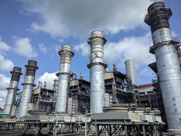 Several large industrial chimneys tower over a complex network of metal structures and machinery, set against a partly cloudy sky.