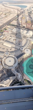 Aerial view overlooking an urban landscape with large geometric buildings, roads, and a visible turquoise water feature. The shadow of a tall structure is cast on the ground, indicating a densely constructed area with modern architectural designs.