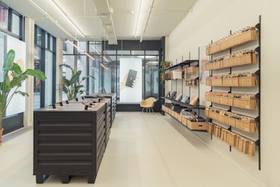 A modern retail space features minimalist design with display shelves along one wall holding neatly organized packages. A central table displays electronic devices on stands. Large windows allow natural light to brighten the space, where potted plants add a touch of greenery.
