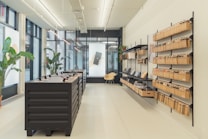 A modern retail space features minimalist design with display shelves along one wall holding neatly organized packages. A central table displays electronic devices on stands. Large windows allow natural light to brighten the space, where potted plants add a touch of greenery.