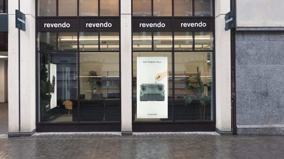 A storefront with large glass windows and multiple signs displaying 'revendo'. Inside, there are visible chairs, plants, and shelves. A poster on the window features the words 'Sell. Repair. Buy.' along with an image of a hand interacting with an electronic device.