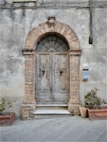 An ancient wooden double door set in a rustic brick archway, with peeling paint and a weathered appearance. Below the door is a stone step, flanked by two rectangular planters containing small plants. A metal mailbox is attached to the wall on the right, and the building facade shows signs of age and wear, with cracks visible in the plaster.