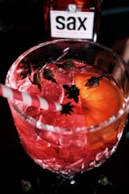 A close-up of a vibrant, red-colored cocktail in a textured glass, garnished with an orange slice, small flower buds, and a red and white striped straw. A bottle with a label reading 'sax' is visible in the background.