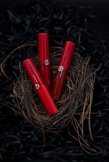 Three red lipstick tubes are elegantly arranged in a nest made of twigs, set against a dark background. The delicate natural materials contrast with the sleek, glossy finish of the lipsticks.