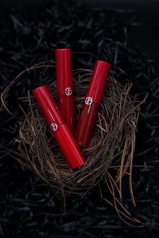 Three red lipstick tubes are elegantly arranged in a nest made of twigs, set against a dark background. The delicate natural materials contrast with the sleek, glossy finish of the lipsticks.