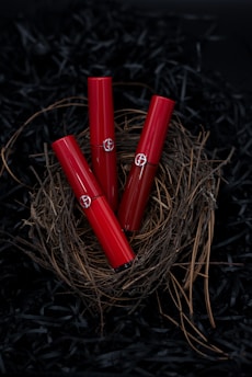 Three red lipstick tubes are elegantly arranged in a nest made of twigs, set against a dark background. The delicate natural materials contrast with the sleek, glossy finish of the lipsticks.