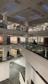 A modern art gallery with multiple levels and open spaces, featuring various artworks on the walls. The structure is illuminated by soft indoor lighting, highlighting paintings and photographs. The gallery features glass railings and a spacious, geometric design.