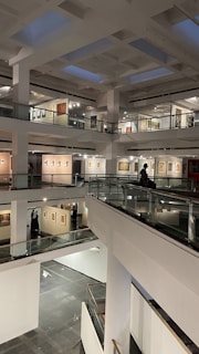 A modern art gallery with multiple levels and open spaces, featuring various artworks on the walls. The structure is illuminated by soft indoor lighting, highlighting paintings and photographs. The gallery features glass railings and a spacious, geometric design.
