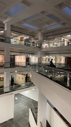 A modern art gallery with multiple levels and open spaces, featuring various artworks on the walls. The structure is illuminated by soft indoor lighting, highlighting paintings and photographs. The gallery features glass railings and a spacious, geometric design.