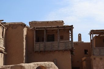 A traditional mud-brick building with wooden balconies and beams, featuring small windows and a flat roof. The structure is set against a clear blue sky, casting shadows that emphasize its rustic texture and earthy tones.