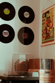 A cozy corner featuring a collection of vinyl records mounted on the wall, with a colorful artwork poster alongside them. Below, there is a turntable with a glass cover, a decorative framed art piece, and a wooden crate filled with vinyl records.