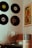 A cozy corner featuring a collection of vinyl records mounted on the wall, with a colorful artwork poster alongside them. Below, there is a turntable with a glass cover, a decorative framed art piece, and a wooden crate filled with vinyl records.