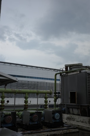 A cloudy sky looms over an industrial setting featuring large machinery and piping. The view includes a gray, metal-clad building in the background, and weathered equipment in the foreground, hinting at an industrial or mechanical environment.