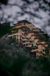A multi-story, modern residential building nestled on a lush, green hillside. The structure features terraces, balconies, and geometric architectural elements, surrounded by dense vegetation and tall trees.