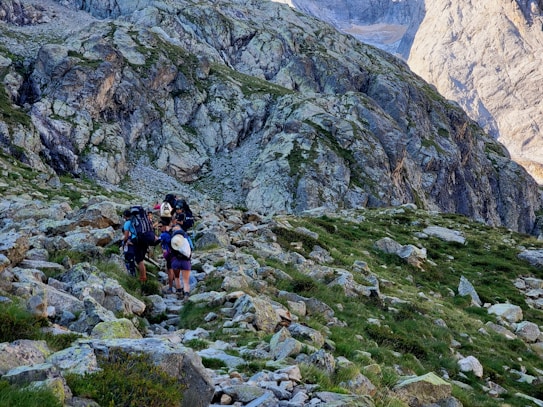 A group of hikers is ascending a rocky mountain trail surrounded by rugged, steep cliffs. The landscape is a mix of gray stones and patches of green grass, indicating an alpine environment. The sky is clear, hinting at a sunny day.