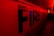 Large capital letters spelling 'FIRE' are prominently featured against a red backdrop. The scene is illuminated with intense red and yellow lighting, creating a dramatic and urgent atmosphere. The setting appears to be indoors, possibly a hallway or tunnel.