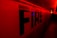 Large capital letters spelling 'FIRE' are prominently featured against a red backdrop. The scene is illuminated with intense red and yellow lighting, creating a dramatic and urgent atmosphere. The setting appears to be indoors, possibly a hallway or tunnel.