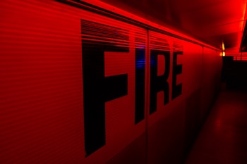 Large capital letters spelling 'FIRE' are prominently featured against a red backdrop. The scene is illuminated with intense red and yellow lighting, creating a dramatic and urgent atmosphere. The setting appears to be indoors, possibly a hallway or tunnel.
