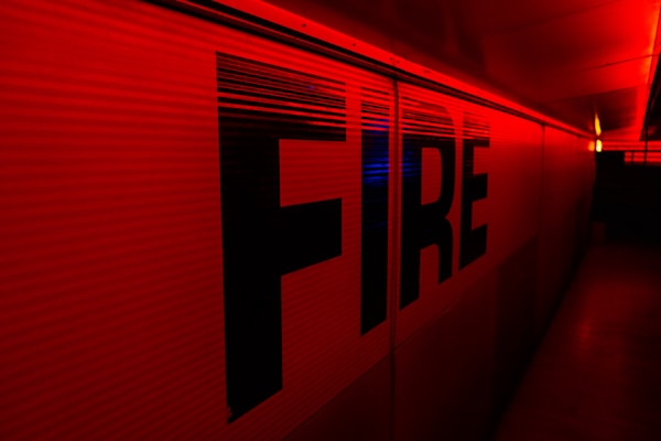 Large capital letters spelling 'FIRE' are prominently featured against a red backdrop. The scene is illuminated with intense red and yellow lighting, creating a dramatic and urgent atmosphere. The setting appears to be indoors, possibly a hallway or tunnel.