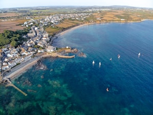 An aerial view of a coastal town with houses clustered near the shore, surrounded by fields and countryside. The coastline forms a gentle curve with rocky outcrops and a sandy beach. Several small sailboats are visible in the clear blue water, scattered throughout the bay.