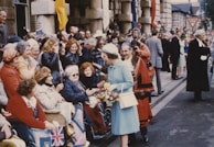 A group of people are gathered on a street during a public event. Many are seated and dressed in colorful attire, waving flags and smiling. The setting appears festive, with individuals engaging with a woman in light blue attire who holds a bouquet. Buildings in the background suggest an urban location.
