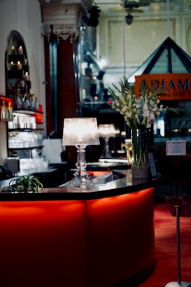 A stylish and elegant bar setting with a curved counter illuminated in red light. A decorative lamp with a clear, ornate base is placed on the counter, casting a soft light. There are tall green plants with white flowers arranged in a vase beside the lamp. A mirrored wall and wooden pillar with intricate carvings add to the sophisticated ambiance. Shelves with bottles and glasses are visible in the background.