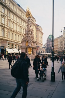 A busy urban street with people walking and a large ornate monument featuring intricate sculptures in the middle. Surrounding the street are historic buildings with classical architecture. The atmosphere is lively with a diverse crowd, including a child in a stroller pushed by an adult.