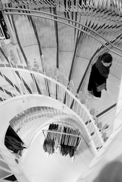 A spiral staircase with elegant railings is viewed from above, showing a person, possibly a woman, walking up or down. A rack of hanging clothes is visible at the bottom of the stairs, suggesting a retail environment.