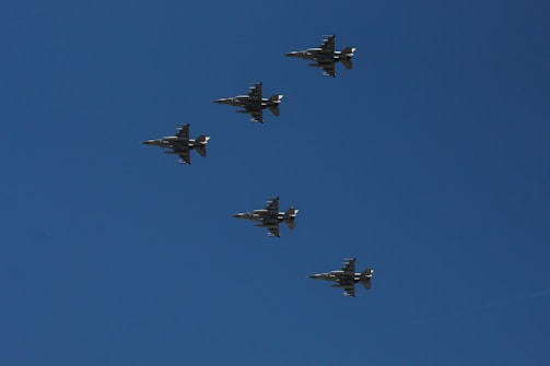 Five military fighter jets fly in a V formation against a clear blue sky.