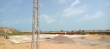 A construction site with a large, flat concrete foundation partially covered by sand and gravel. There is a metal pole or tower on the left side of the image. In the background, there are small hills and scattered buildings. The sky is clear with a few clouds.