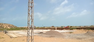 A construction site with a large, flat concrete foundation partially covered by sand and gravel. There is a metal pole or tower on the left side of the image. In the background, there are small hills and scattered buildings. The sky is clear with a few clouds.