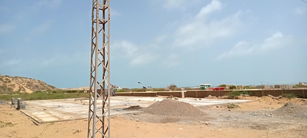 A construction site with a large, flat concrete foundation partially covered by sand and gravel. There is a metal pole or tower on the left side of the image. In the background, there are small hills and scattered buildings. The sky is clear with a few clouds.