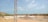 A construction site with a large, flat concrete foundation partially covered by sand and gravel. There is a metal pole or tower on the left side of the image. In the background, there are small hills and scattered buildings. The sky is clear with a few clouds.