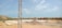A construction site with a large, flat concrete foundation partially covered by sand and gravel. There is a metal pole or tower on the left side of the image. In the background, there are small hills and scattered buildings. The sky is clear with a few clouds.