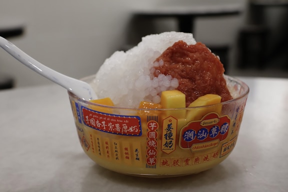 A bowl of dessert featuring a mix of crushed ice, diced mango, red bean paste, and clear tapioca pearls. The bowl is labeled with text and a plastic spoon is resting inside it. The background is blurred, highlighting the dessert.
