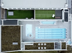 An aerial view of a modern rooftop facility featuring a large swimming pool with multiple lanes, adjacent to a smaller pool or water feature. The rooftop has landscaped grass areas with what appears to be a tennis court setup on one side, and sections covered with foliage, possibly for insulation or eco-friendly purposes. The design is geometric, with clean lines and a mix of concrete and greenery.