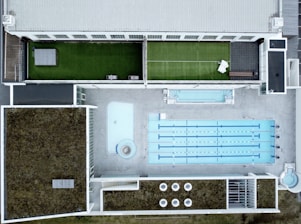 An aerial view of a modern rooftop facility featuring a large swimming pool with multiple lanes, adjacent to a smaller pool or water feature. The rooftop has landscaped grass areas with what appears to be a tennis court setup on one side, and sections covered with foliage, possibly for insulation or eco-friendly purposes. The design is geometric, with clean lines and a mix of concrete and greenery.